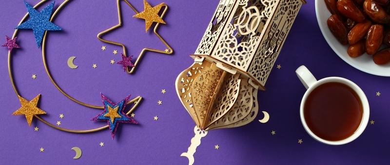 Gold metal wire crescent moon and star with cut-out sparkly blue, purple and gold star details beside gold Ramadan lantern, white mug of brown liquid and white bowl of dates against a purple background.
