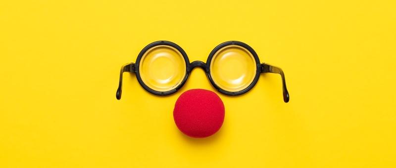 Red clown nose beneath round, black-rimmed comedy glasses against a yellow background.