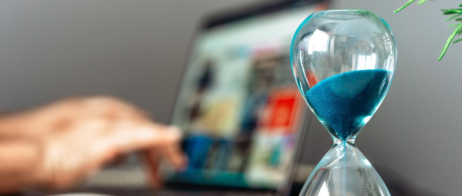 Reloj de arena de cristal con arena azul junto a un ordenador portátil en uso, simbolizando la gestión del tiempo en el trabajo digital.