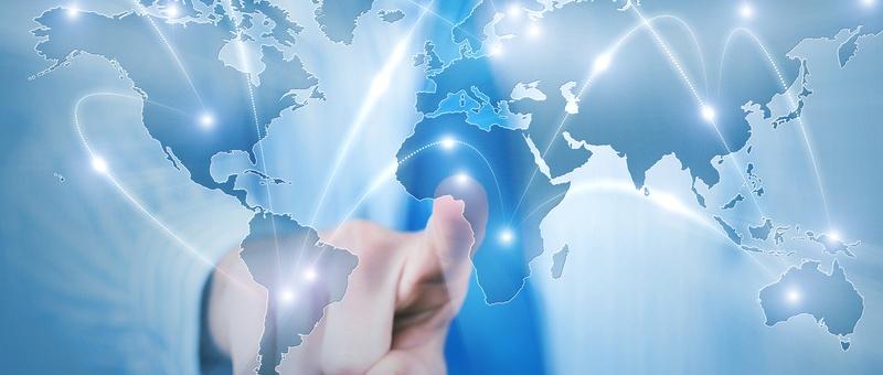 Mano tocando un mapamundi digital con líneas de conexión resplandecientes que unen los continentes sobre un fondo azul.
