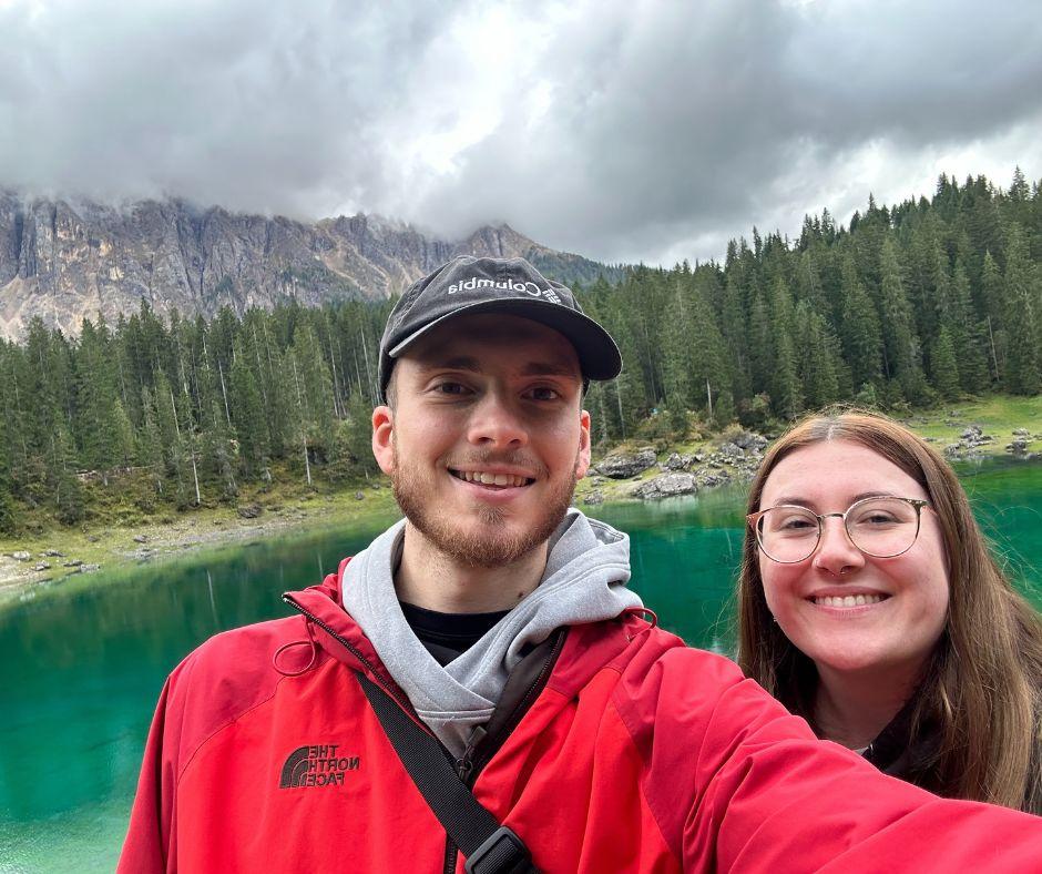Pareja joven posando para un selfie frente a un lago arbolado bajo una cadena montañosa.