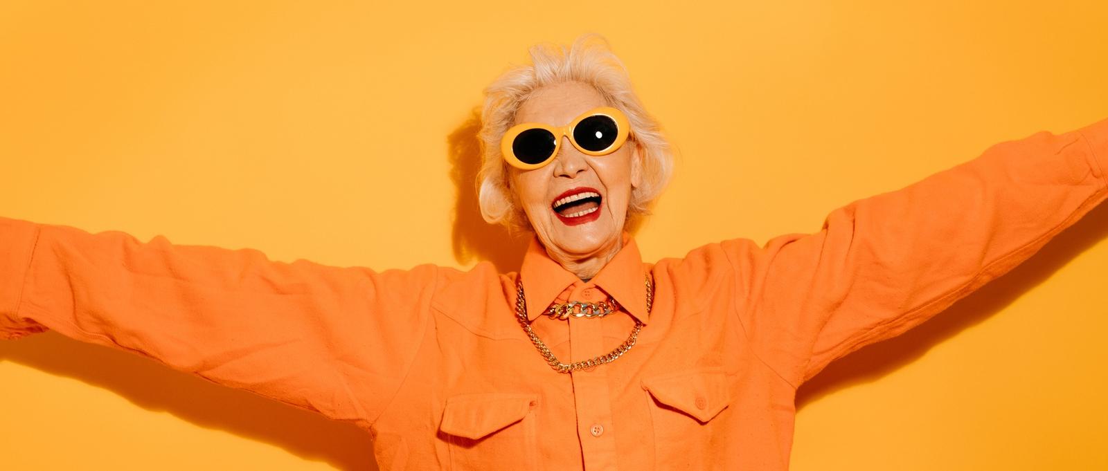 Sonriente mujer elegante de alto nivel con gafas de sol de moda extiende las manos, quiere abrazar a alguien, contento de verte aislados sobre fondo amarillo.