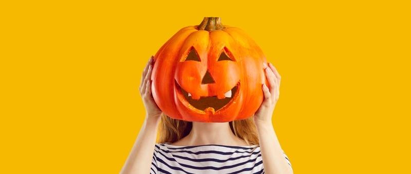 Mujer con top de rayas sobre fondo amarillo escondiendo la cara detrás de un farol de calabaza de Halloween.