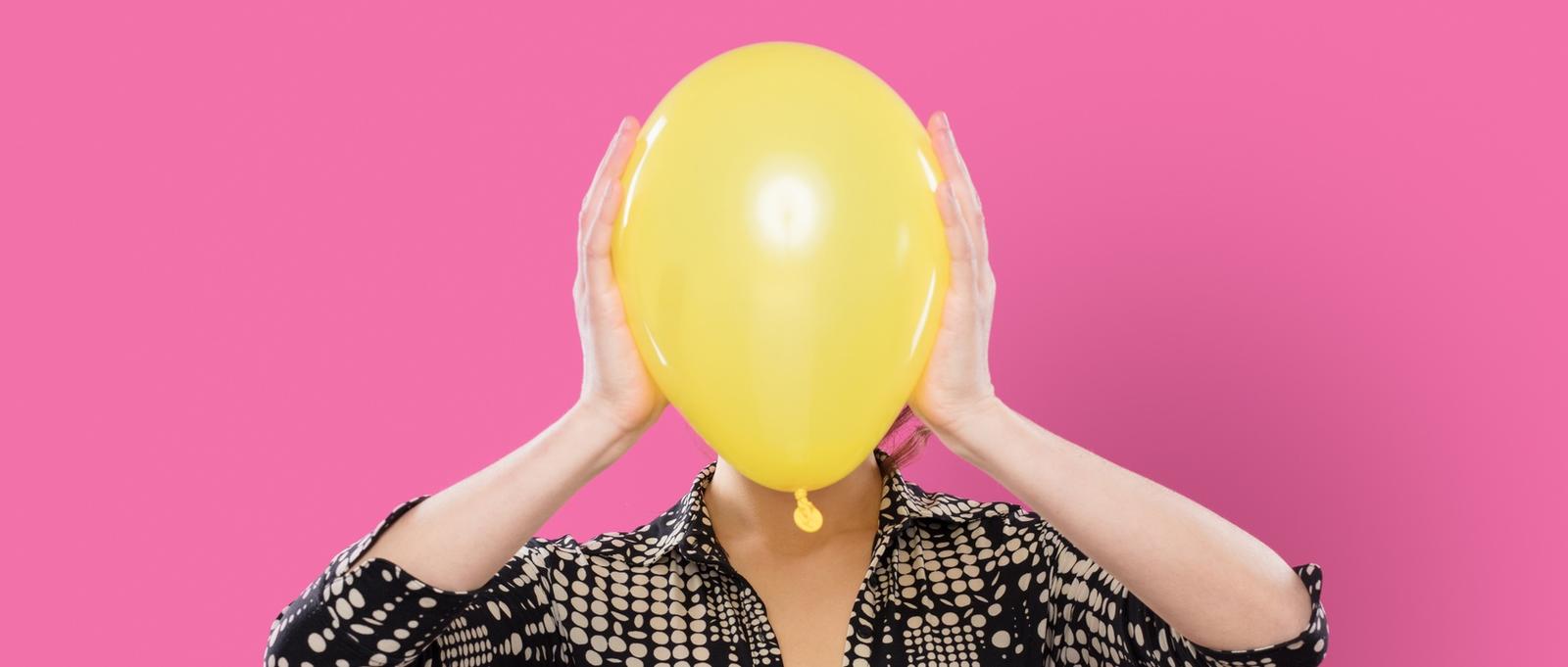 Mujer sobre fondo rosa sosteniendo globo amarillo sobre la cara.