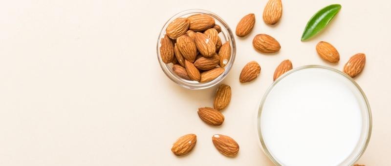 Glass of almond milk beside scattered pile of almond nuts with green leaf garnish against pale beige background.