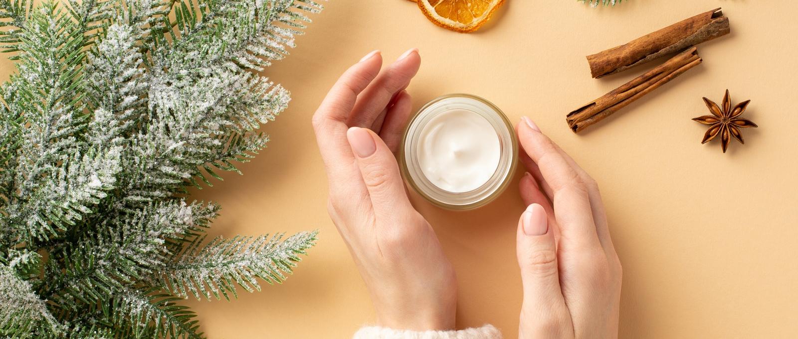 Concepto de cosméticos para el cuidado de la piel en invierno. Primera persona foto de vista superior de las manos de la niña en el tarro de crema jersey ramas de abeto rodajas de naranja seca palitos de canela y anís sobre fondo beige pastel aislado.