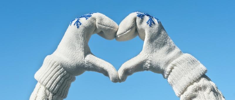 Persona haciendo manos de corazón con jersey blanco de punto y guantes blancos de punto con estampado de copos de nieve, contra un cielo azul invernal.