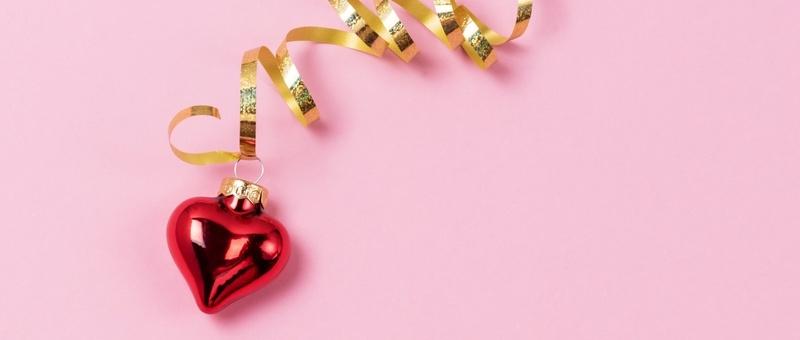 Red, reflective, heart-shaped Christmas tree ornament with shiny gold ribbon against a pink background.