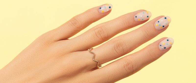 Mano con nail art de lunares de colores sobre esmalte nude que lleva un delicado anillo dorado en zigzag sobre fondo amarillo.
