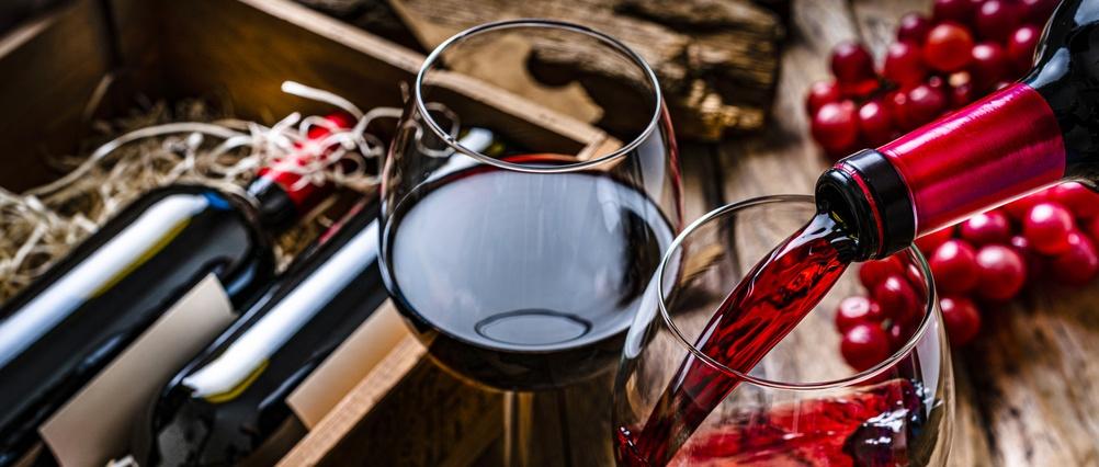 Vino tinto siendo vertido en un vaso junto a una copa llena de vino tinto, con botellas y uvas rojas frescas sobre superficie de madera.