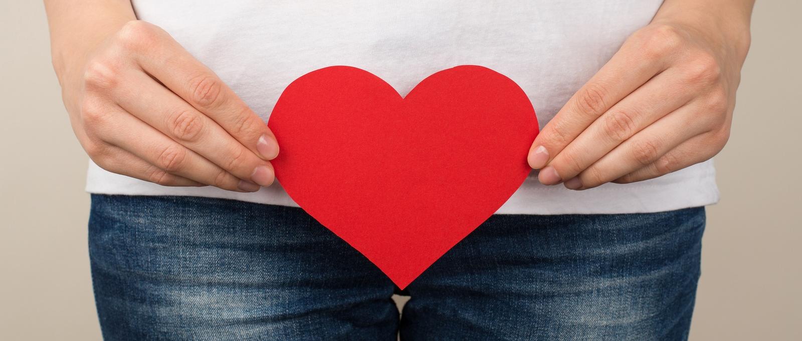 Cropped closeup photo of woman in white t-shirt and jeans holding red paper heart near crotch on isolated grey background
