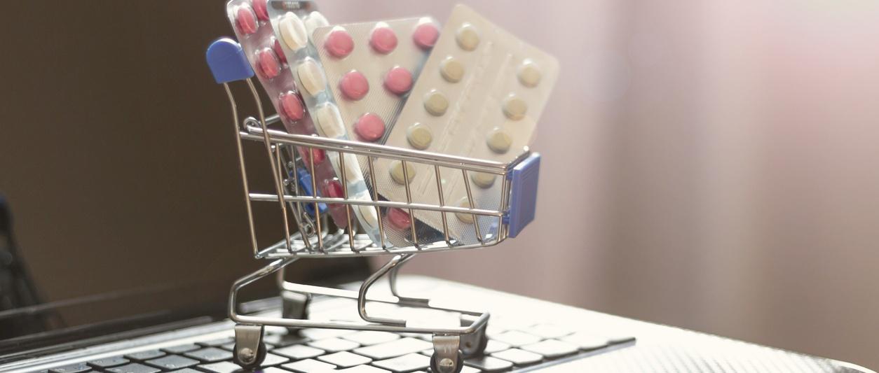 Carrito de la compra en miniatura lleno de blísteres de pastillas anticonceptivas rosas y blancas, sentado sobre el teclado de un ordenador portátil.