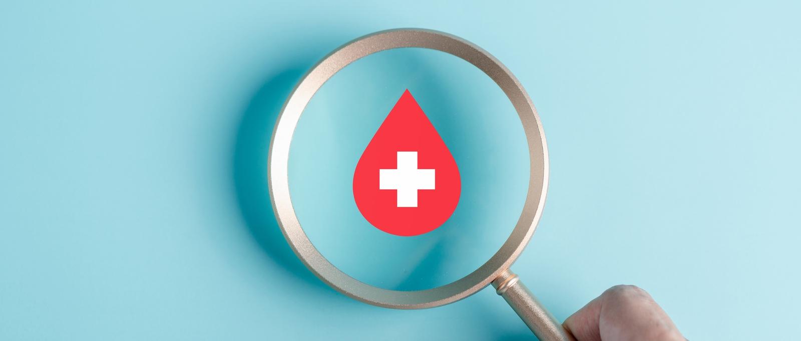 Mano sosteniendo lupa enfocando icono de gota de sangre roja sobre fondo azul pálido.