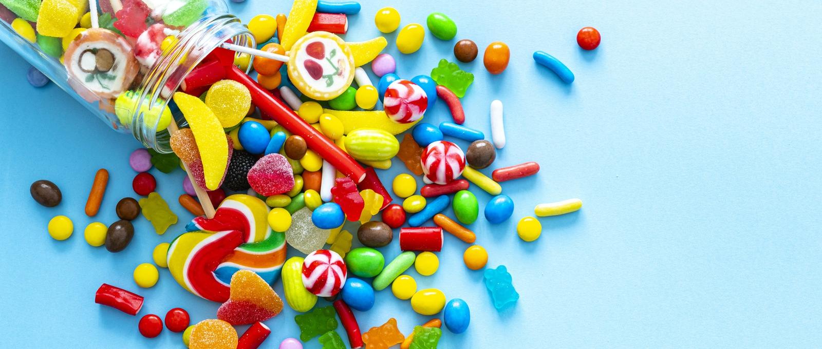 Gran grupo de todo tipo de dulces multicolores derramando de un frasco de vidrio tiro desde arriba sobre fondo azul.