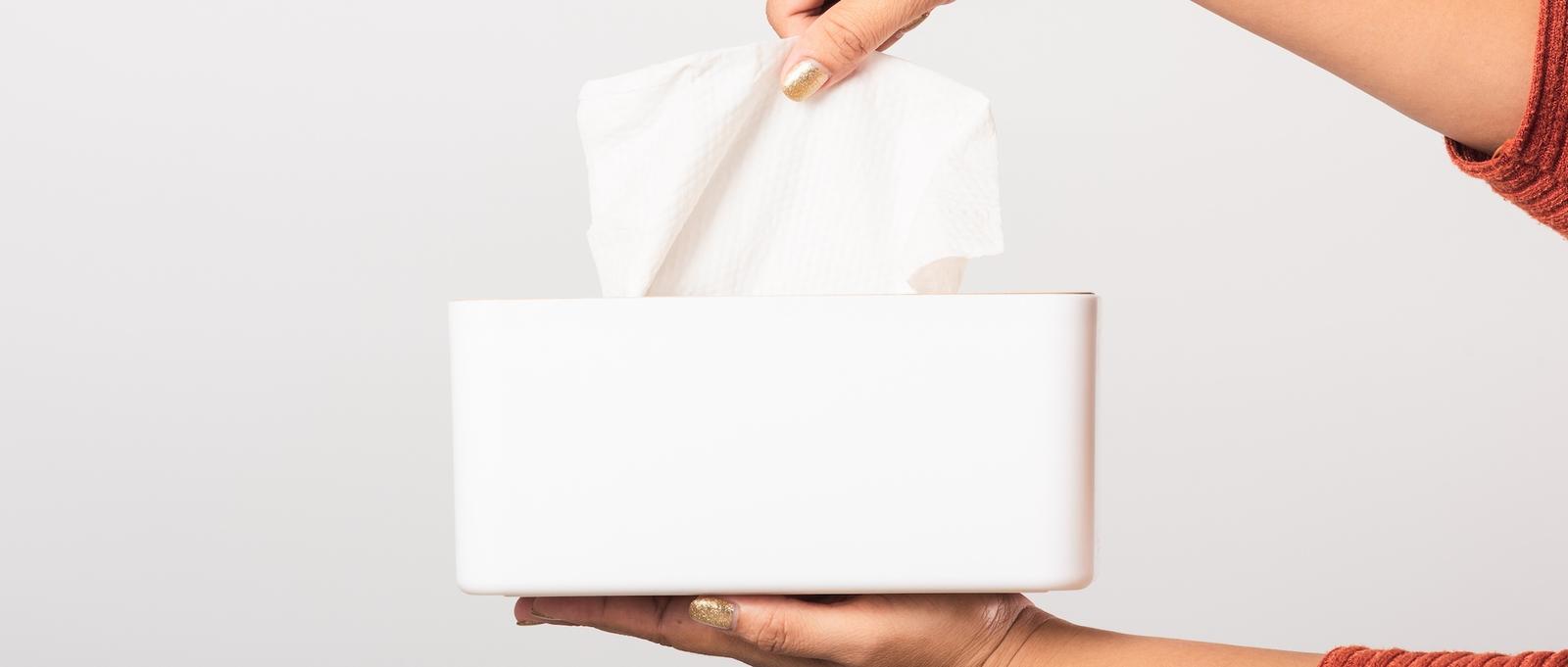 Mano con esmalte de uñas dorado sacando un pañuelo de una caja rectangular blanca sobre fondo gris claro.