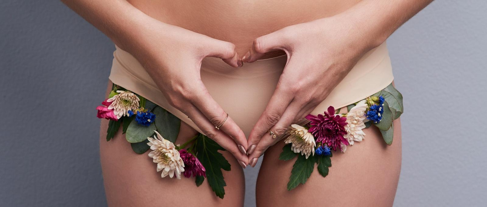 Foto de estudio recortada de una mujer con ropa interior de flores y haciendo un gesto en forma de corazón sobre un fondo gris.