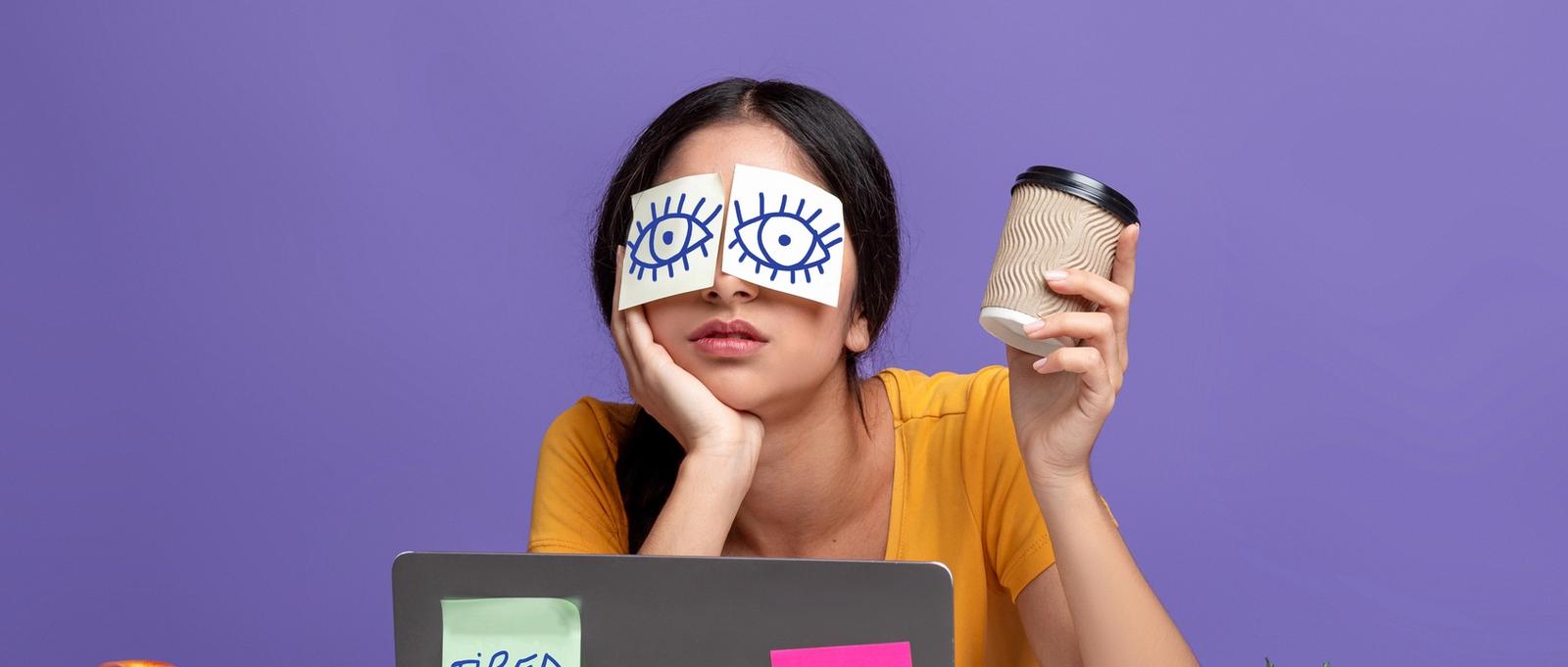Retrato de mujer joven exhausta sentada en su lugar de trabajo con pegatinas en los ojos y sosteniendo una taza con café, trabajando en un ordenador portátil en una mesa sobre un fondo de estudio morado.