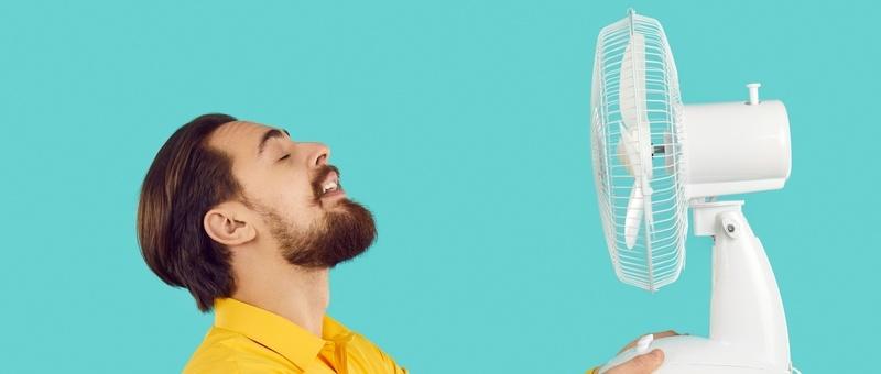 Hombre barbudo divertido disfrutando de la brisa de un ventilador eléctrico sobre un fondo azul.