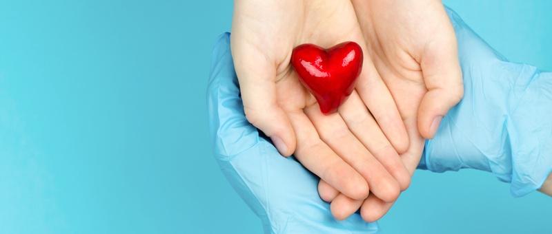 Paciente y profesional sanitario con las manos enguantadas sosteniendo juntos un corazón rojo brillante sobre fondo azul.