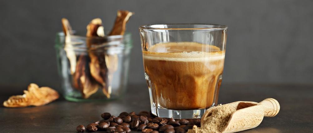 Café con setas en un vaso, granos de café y setas secas sobre fondo oscuro.