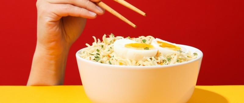 Isolated hand holding wooden chopsticks, appearing from behind yellow table against red background, preparing to tuck in to bowl of noodles and egg.