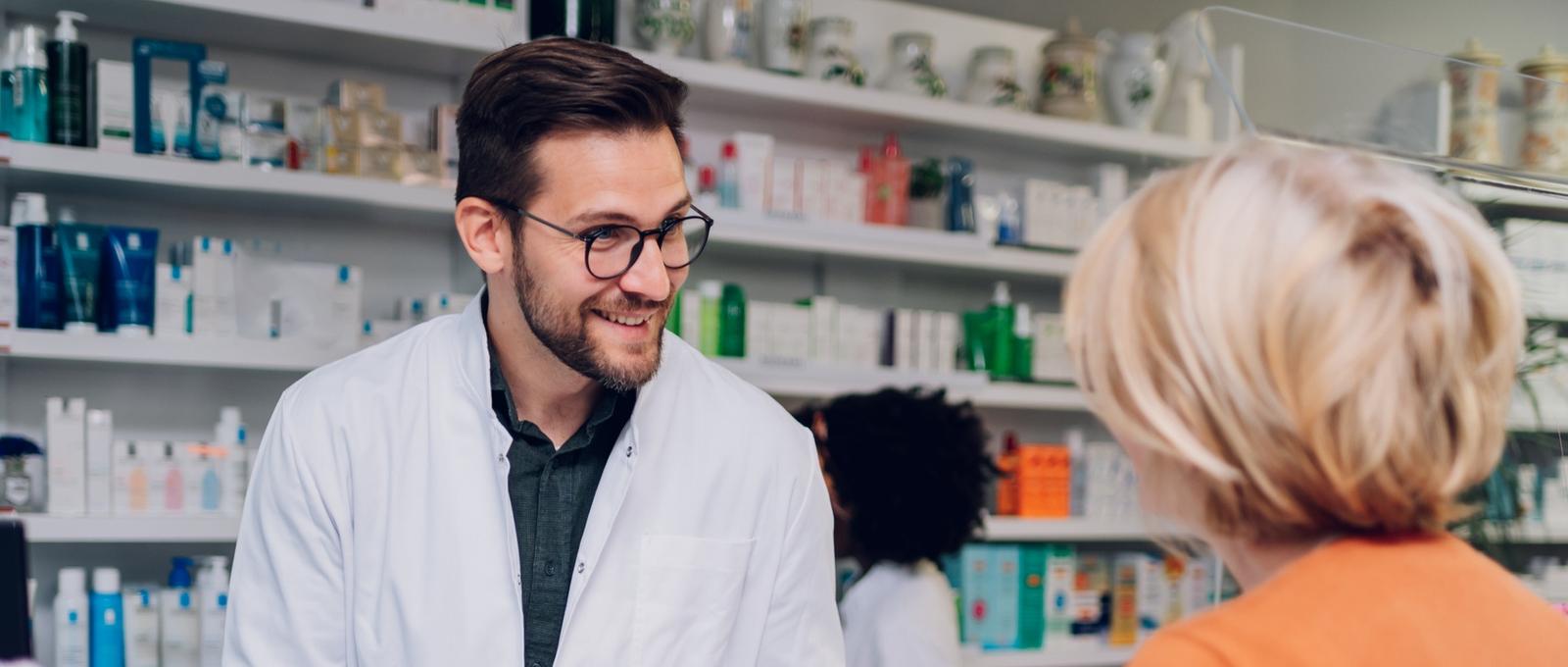 Joven farmacéutico hablando con una clienta en una farmacia con una farmacéutica al fondo. Estantes con productos para el cuidado de la salud.
