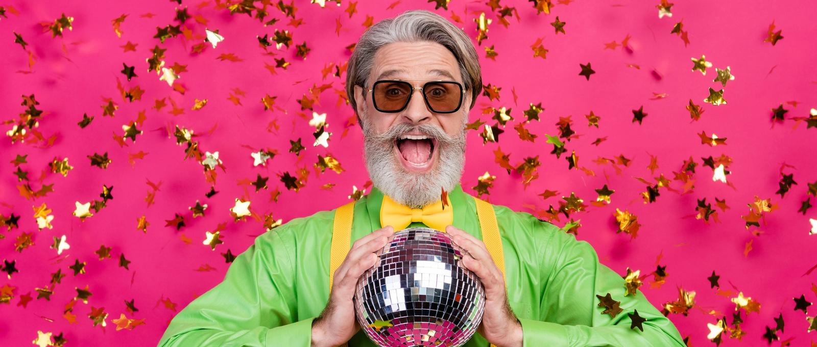 Foto de divertido abuelo fiesta club confeti caída emocionado boca abierta sostener brillante bola de discoteca llevar gafas de sol camisa verde tirantes amarillos pajarita, aislado brillo fondo de color rosa