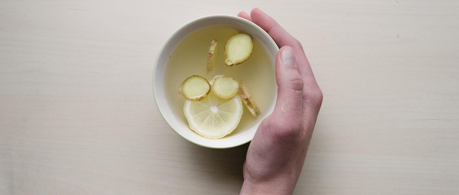 Mano sosteniendo un cuenco blanco de té de jengibre y limón con ingredientes troceados flotando en un líquido transparente.