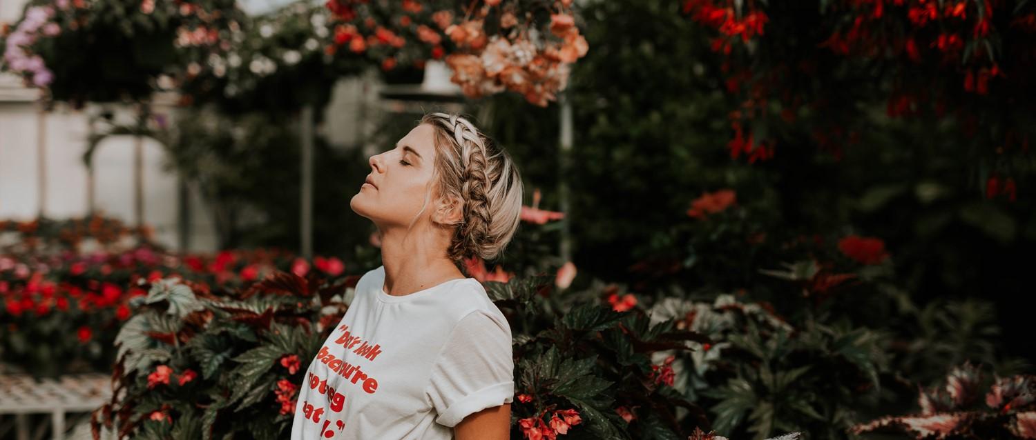 Mujer con el pelo rubio trenzado en camiseta blanca rodeada de flores de color rojo vibrante y exuberante vegetación en un jardín.