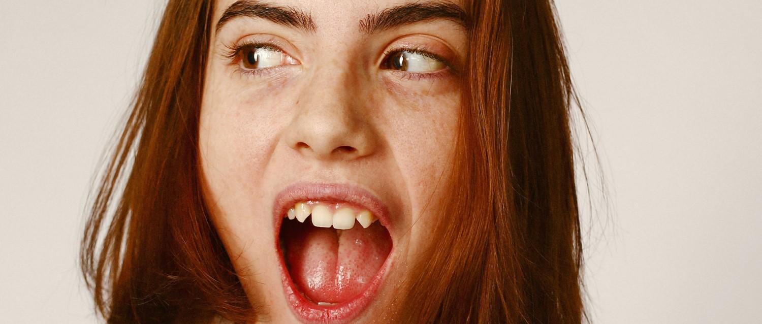 Retrato en primer plano con pelo castaño que muestra una boca abierta y ojos que miran de reojo sobre un fondo neutro.