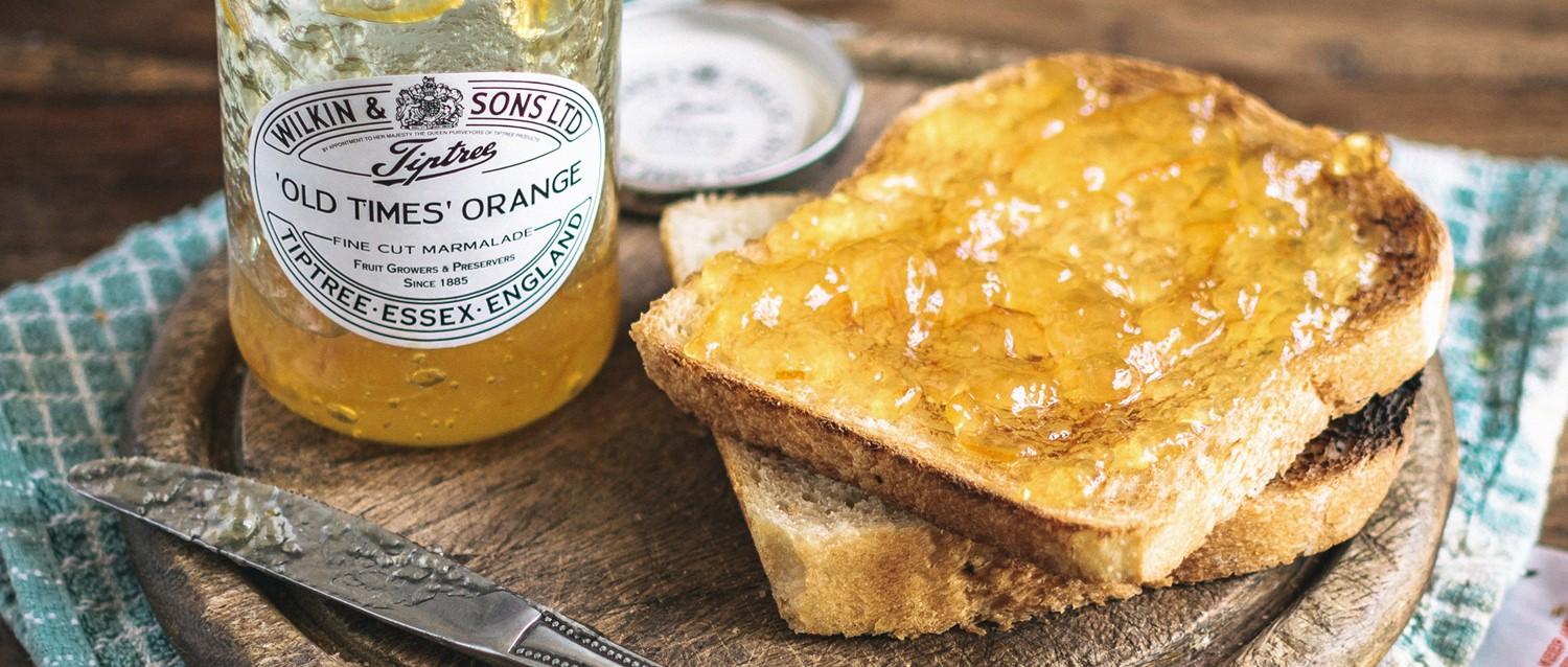 Tostada untada con mermelada de naranja Wilkin & Sons 'Old Times' sobre tabla de madera con tarro y cuchillo sobre paño verde azulado.