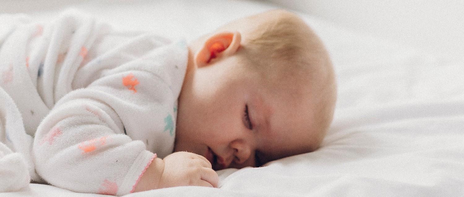 Bebé dormido en ropa de dormir blanca con dibujos descansando plácidamente sobre ropa de cama blanca, con una manita enroscada cerca de la cara.