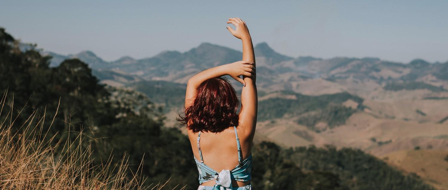 Persona de pelo castaño levantando el brazo hacia el paisaje montañoso, vista de espaldas con atuendo veraniego.