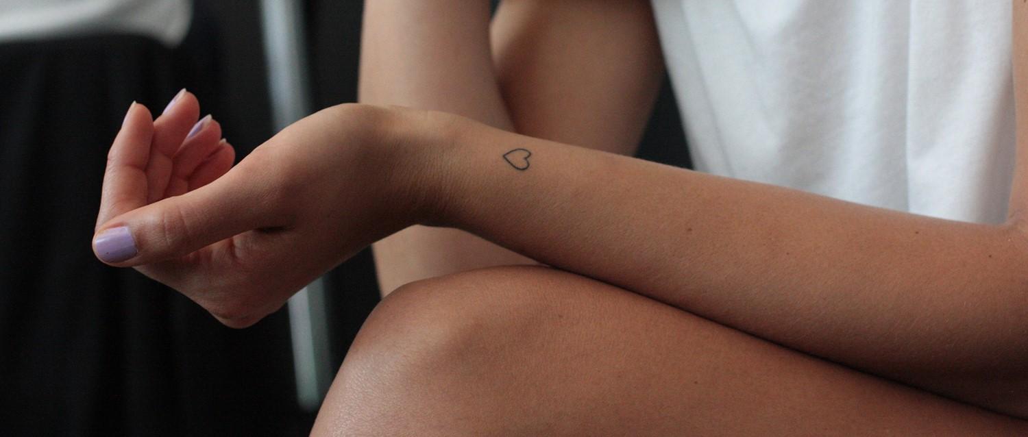 Primer plano de la muñeca de una persona con un pequeño tatuaje de corazón, mostrando el esmalte de uñas púrpura pálido y la posición relajada de la mano.