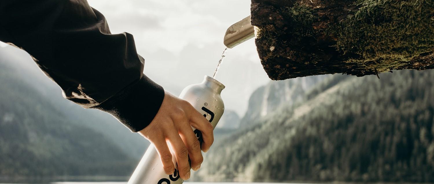 Persona llenando una botella de agua reutilizable de un manantial natural en un paisaje montañoso con espectaculares picos rocosos de fondo.