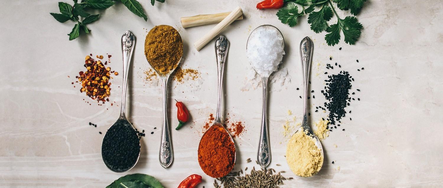 Cucharas de plata con coloridas especias como curry, pimentón, sal y semillas negras, rodeadas de hierbas frescas y guindillas.