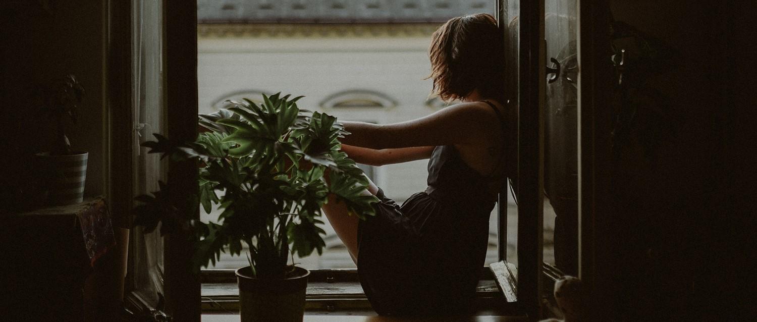 Persona sentada a la tenue luz de la ventana, cuidando de las plantas de interior, creando una atmósfera tranquila y contemplativa.