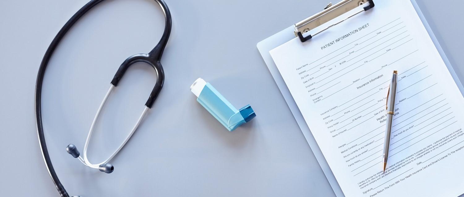 Estetoscopio, inhalador azul y formulario de información del paciente con bolígrafo sobre superficie azul claro, representando la atención médica.