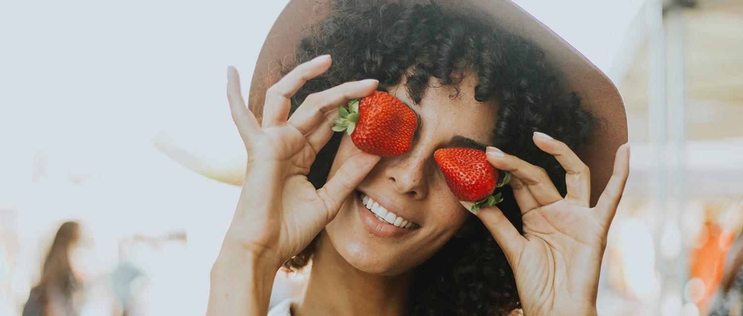 Person with curly hair holding fresh strawberries over their eyes, smiling playfully in soft natural light.