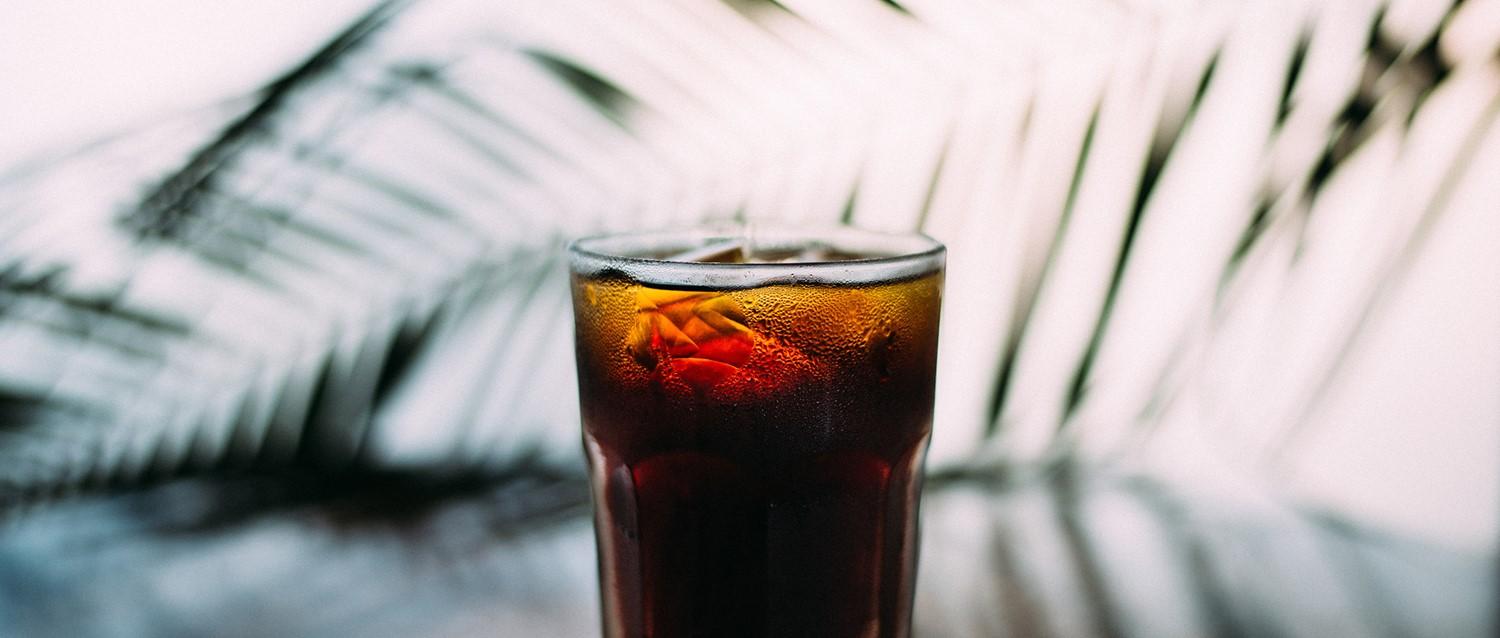 Vaso frío de cola con cubitos de hielo y condensación sobre un fondo difuminado con sombras de hojas de palmera.