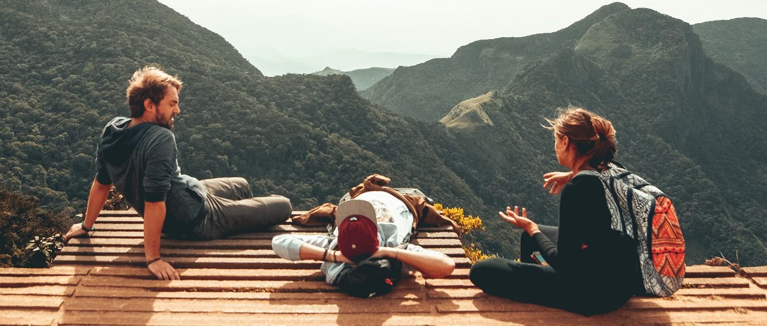 Senderistas descansando en una plataforma de madera con vistas a un espectacular paisaje montañoso con frondosos valles verdes y picos lejanos.