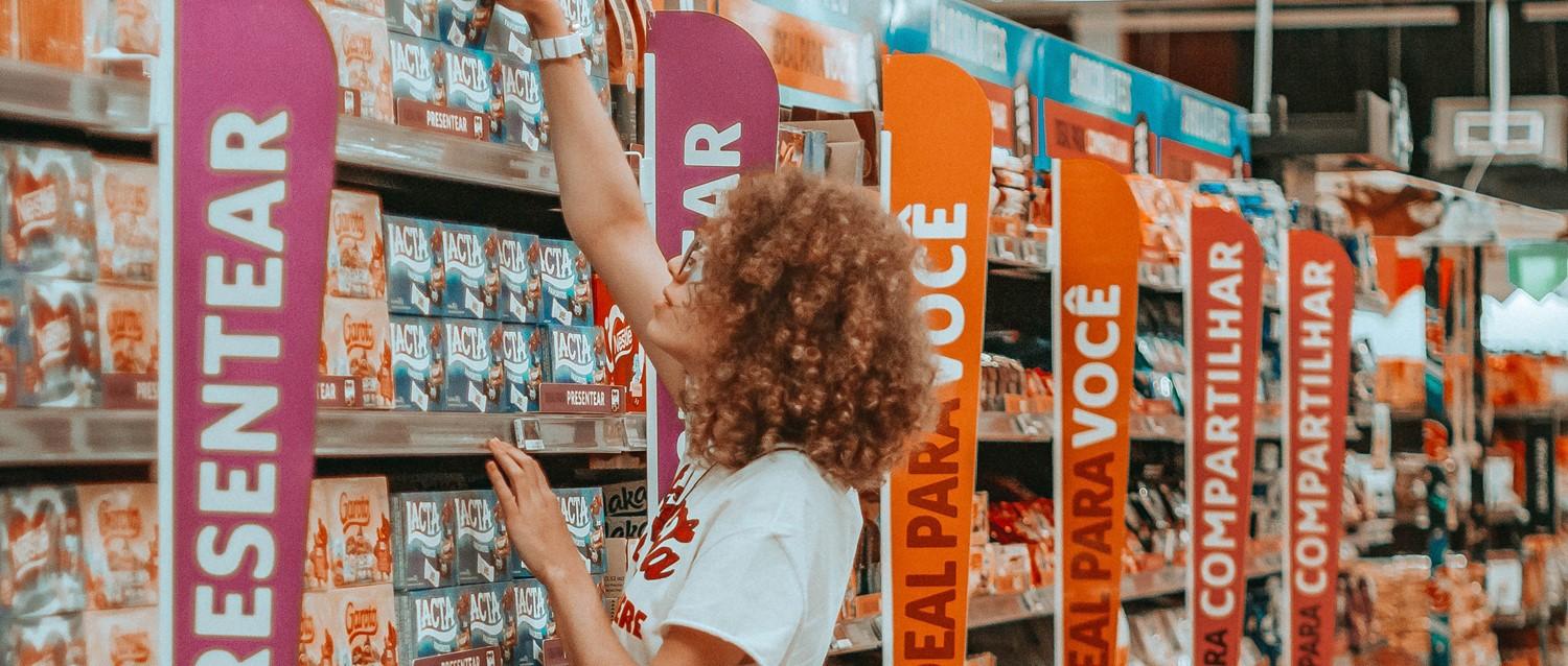 Persona con el pelo rizado buscando productos en un pasillo de supermercado con coloridos carteles de categorías en rosa y naranja.