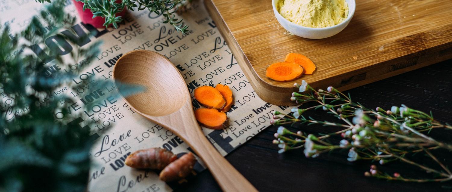 Wooden spoon on "love" patterned cloth with carrot slices, yellow spice in bowl, herbs and flowers for cooking.