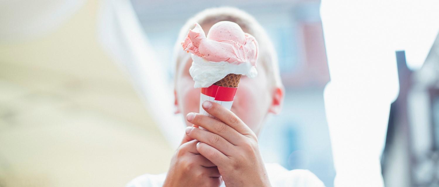 Manos sosteniendo un cucurucho de helado de fresa con bolas rosas y nata montada sobre un fondo brillante y borroso.