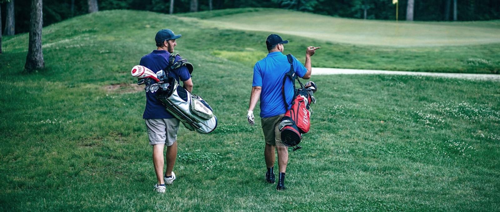 Dos golfistas caminan por un campo verde cargados con bolsas de golf, una plateada y otra roja, en dirección al siguiente hoyo.