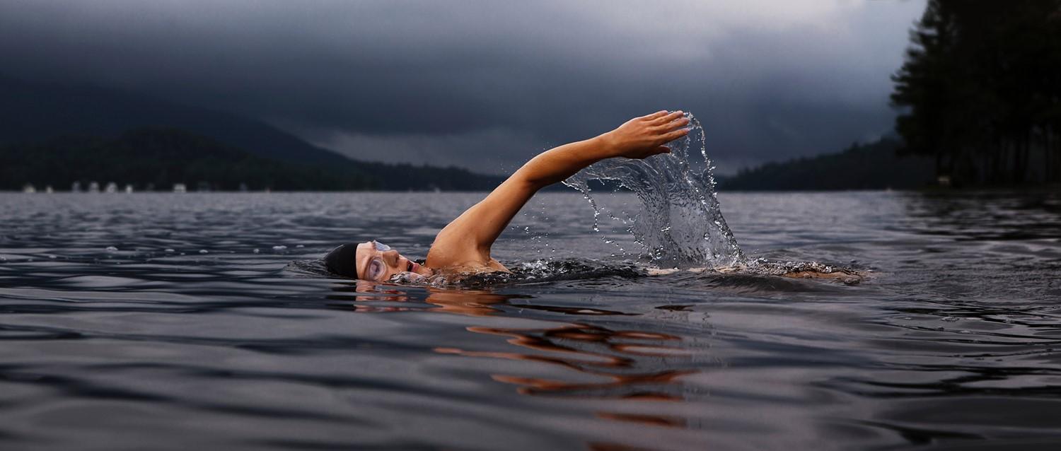 Nadador haciendo crawl frontal en un lago de montaña brumoso, brazo creando salpicaduras contra fondo oscuro cielo tormentoso.
