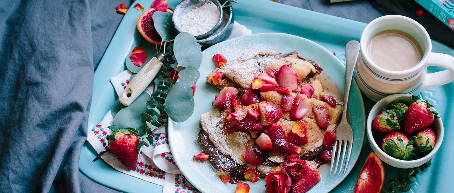 Bandeja de desayuno con crepes cubiertos con fresas frescas, azúcar en polvo, café y un pequeño bol de fruta.