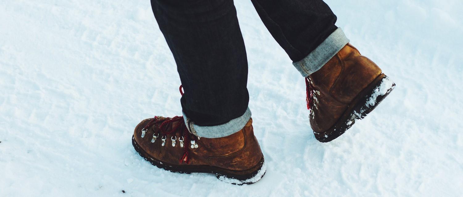 Persona con botas de montaña marrones con cordones rojos y vaqueros oscuros arremangados caminando por la nieve.