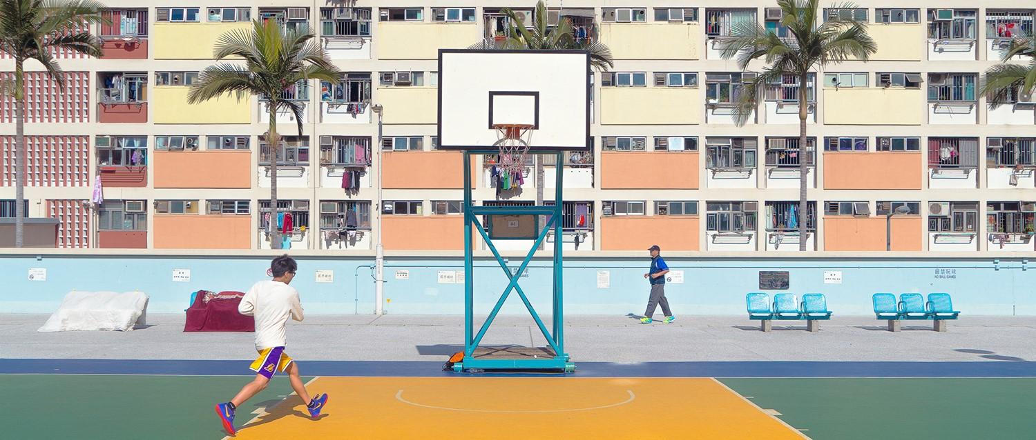 Cancha de baloncesto con un colorido edificio de apartamentos de fondo, palmeras y gente caminando por la cancha.