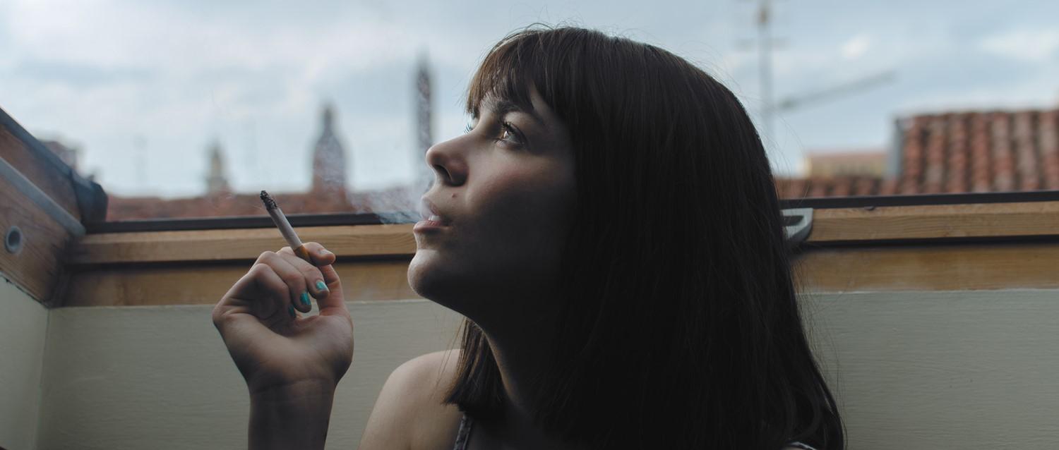 Persona de pelo oscuro fumando junto a una ventana, mirando pensativo el horizonte de la ciudad con el esmalte de uñas verde azulado a la vista.